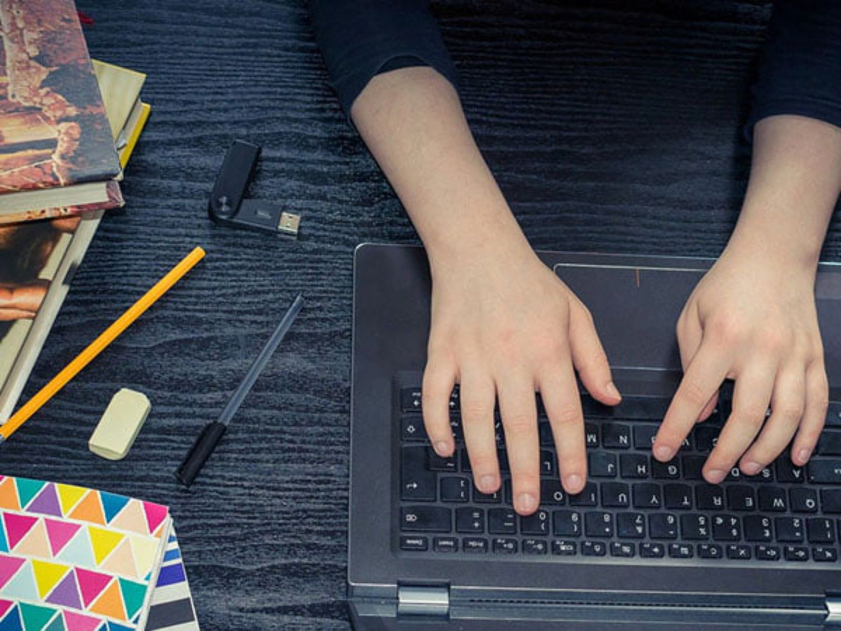 Mani intente a lavorare sulla tastiera di un laptop con attorno strumenti di lavoro (cancelleria, quaderni, ecc)